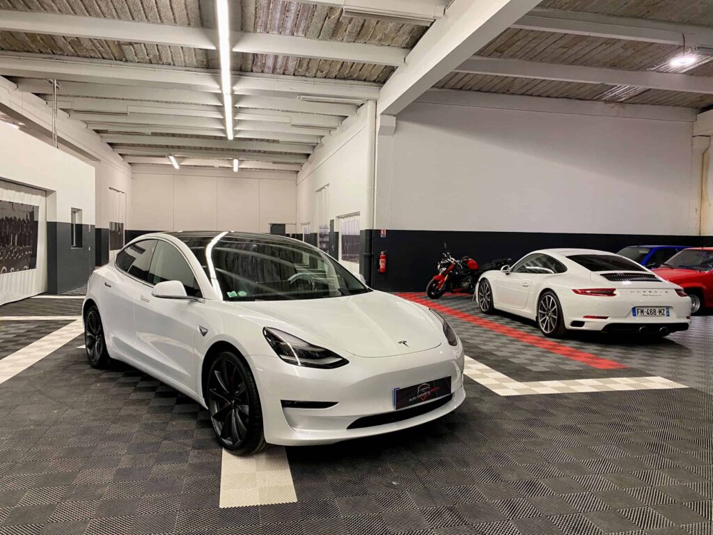 Photo du garage Auto Detailing Reims montrant plusieurs véhicules, dont une Tesla au premier plan, et une Porsche à côté.