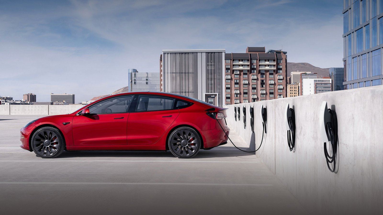 Tesla Model 3 rouge branchée à une rangée de bornes de recharge murales sur un parking en toiture