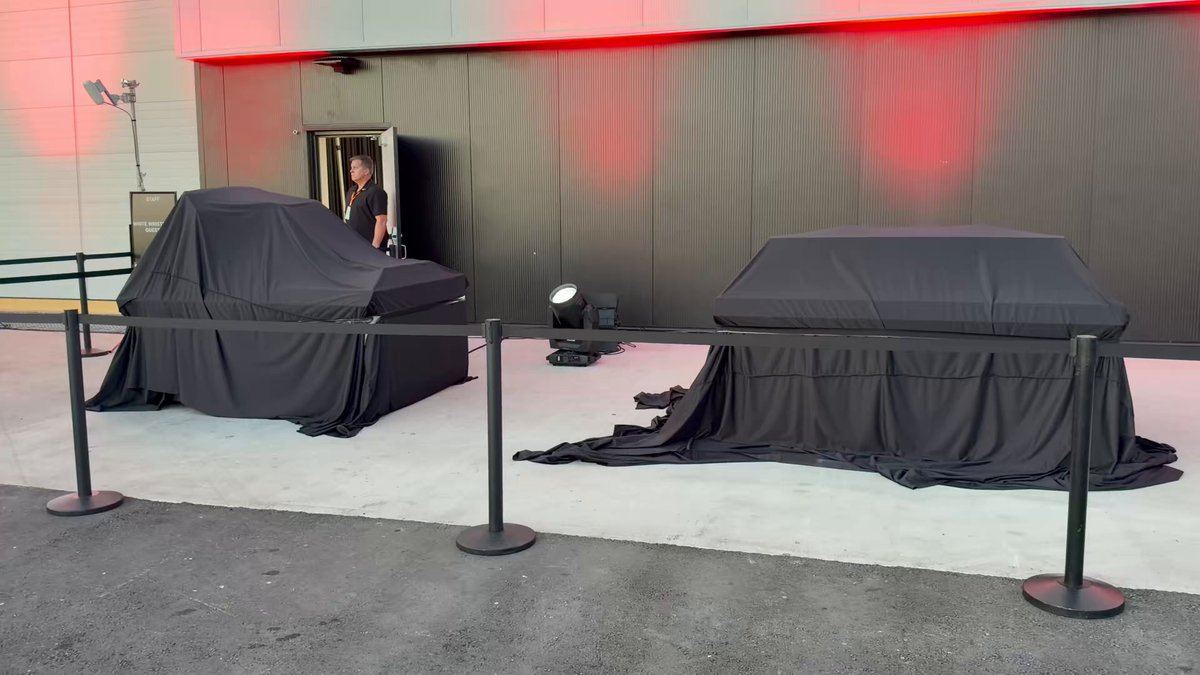 Deux véhicules couverts de bâches noires devant un bâtiment éclairé en rouge sur le site de la présentation du Tesla Roadster